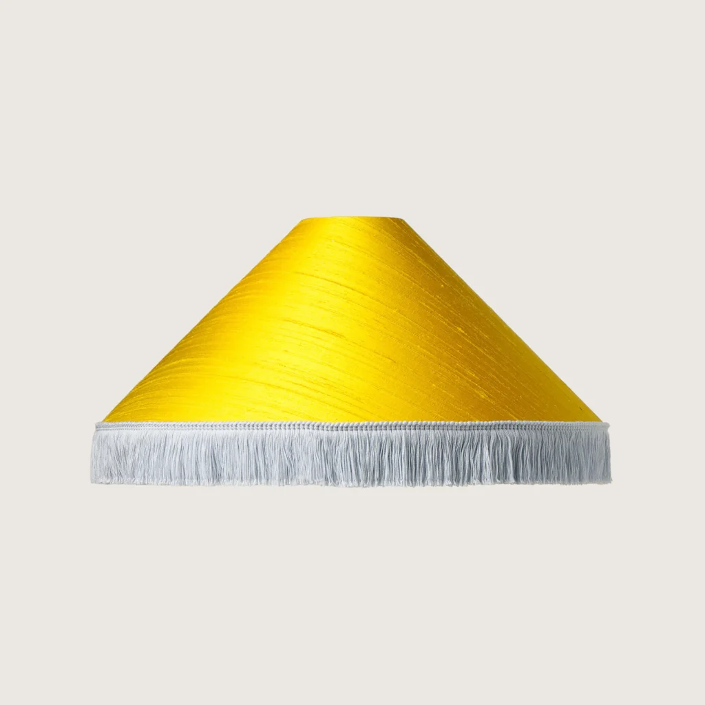 A beautifully formed yellow conical lampshade, finished with a delicate off-white fringe along the bottom edge.