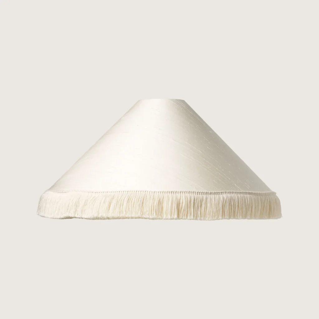 A beautifully formed white conical lampshade, finished with a delicate white fringe along the bottom edge.